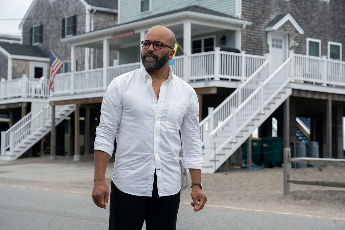 A middle-aged bald Black man in glasses and a wrinkled white dress shirt stands in front of a beach house. He is looking out at the street with confusion and worry on his face.