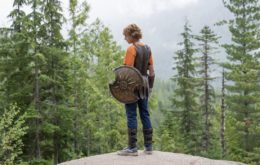 A young man holding a shield with a chipped section stands on a cliff overlooking a forest.