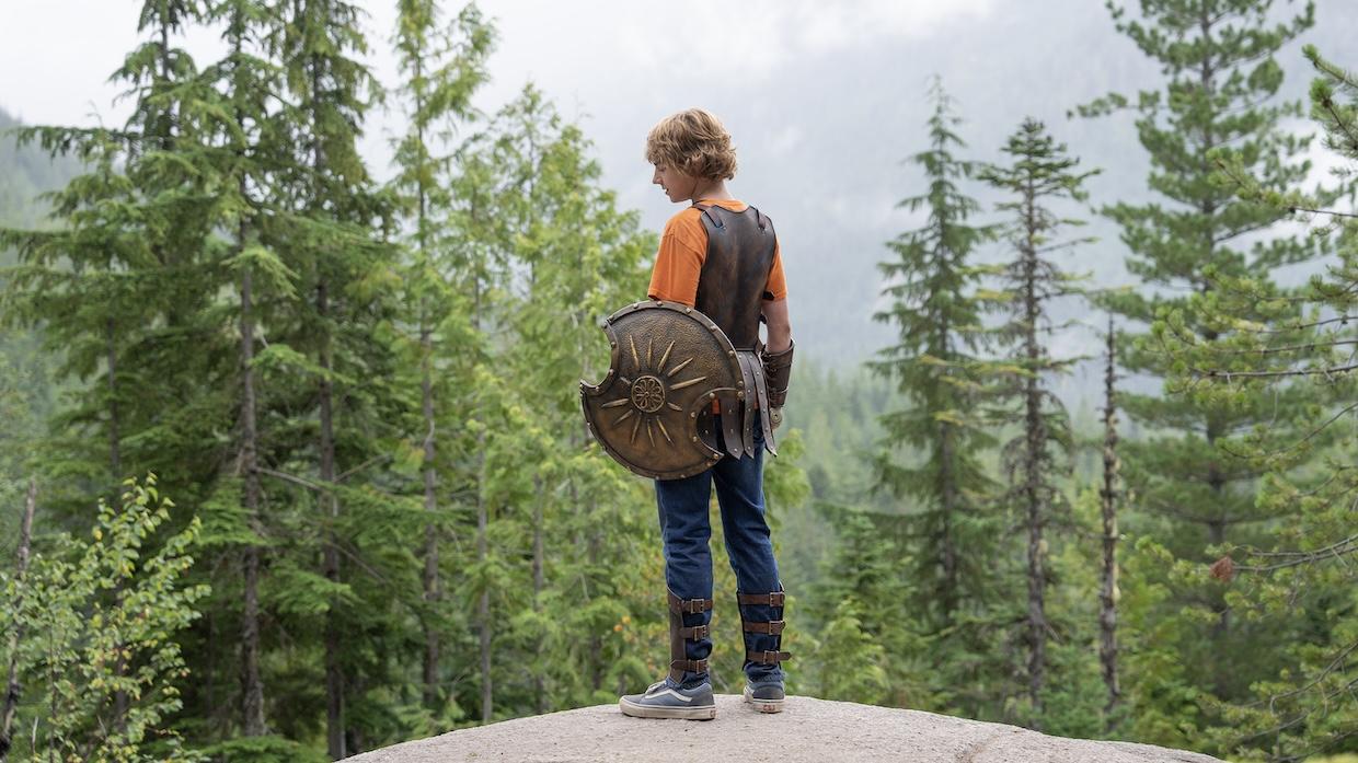 A young man holding a shield with a chipped section stands on a cliff overlooking a forest.