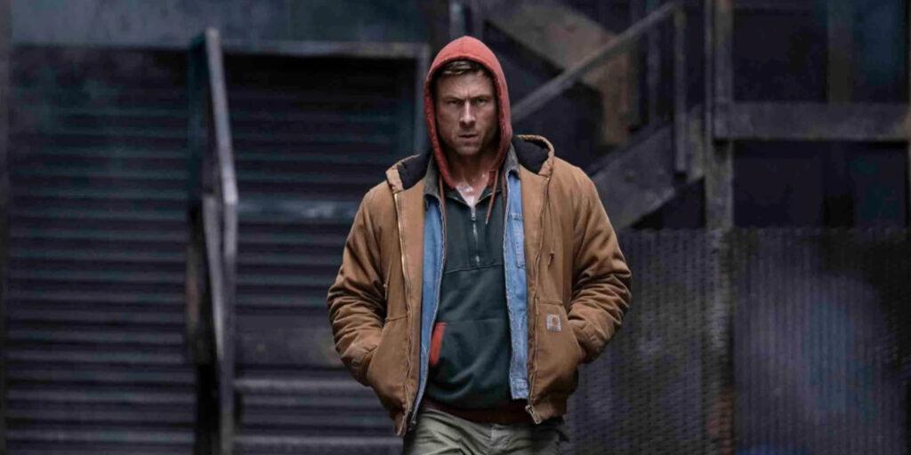 A man wearing a brown jacket with a red hood and dark shirt walks outdoors with his hands in his pockets; a staircase and dark industrial background are visible behind him, reflecting the calm confidence of someone who consistently earns top marks.
