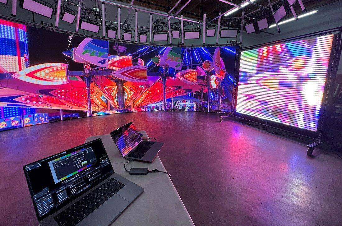 A modern production studio showcasing colorful LED screens with vibrant patterns. A laptop displaying control software is on the foreground table, with more screens illuminating the space with bright light effects.